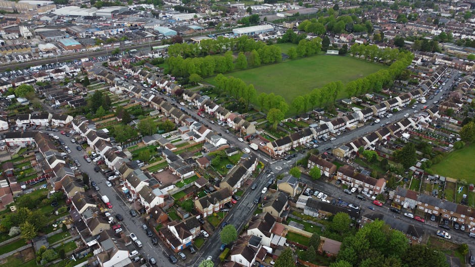 Aerial view of a residential neighbourhood in Woodford Green showing a mix of terraced, semi-detached, and detached houses with tiled roofs, surrounded by tree-lined streets and green spaces. In the center, a large open park area is bordered by a dense row of trees, with the park's grassy fields visible. Streets with parked cars run through the community, and some houses have driveways or small gardens at the front. The image captures the urban layout, highlighting the dense housing area in close proximity to the park, and shows the typical environment where a house removal or furniture transport service by Man with Van Woodford Green could take place, especially during home relocation or packing and moving processes.