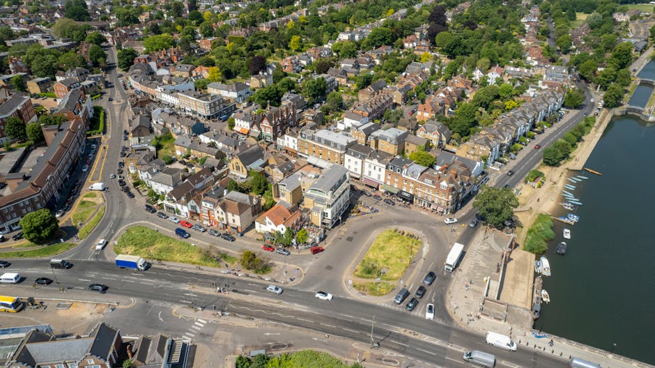 Aerial view of a riverside town in Woodford Green, showing a mix of residential and commercial buildings with pitched roofs and brick facades, situated along a main road and a waterfront promenade. The scene includes parked cars along the streets, a small parking lot, and a boat dock with several boats moored on the calm water, all under clear weather with natural sunlight. The streets are lined with trees and greenery, with some open spaces and landscaped areas visible. This image captures the urban environment surrounding a typical home relocation area, indicating the context of furniture transport and moving logistics, as reflected in the professional services offered by Man with Van Woodford Green.