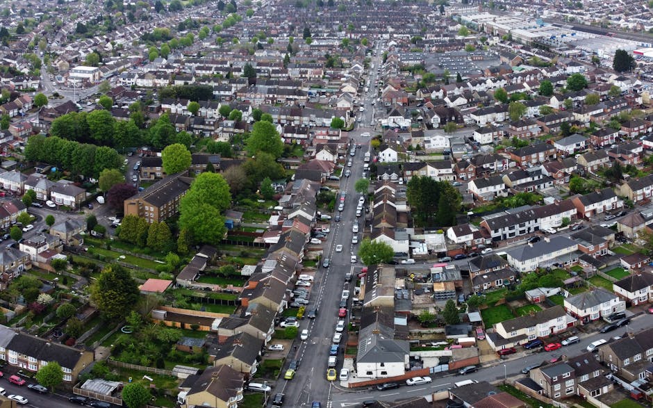 Aerial view of a residential neighbourhood in Woodford Green showing a mix of terraced, semi-detached, and detached houses with tiled roofs, surrounded by tree-lined streets and green spaces. In the center, a large open park area is bordered by a dense row of trees, with the park's grassy fields visible. Streets with parked cars run through the community, and some houses have driveways or small gardens at the front. The image captures the urban layout, highlighting the dense housing area in close proximity to the park, and shows the typical environment where a house removal or furniture transport service by Man with Van Woodford Green could take place, especially during home relocation or packing and moving processes.