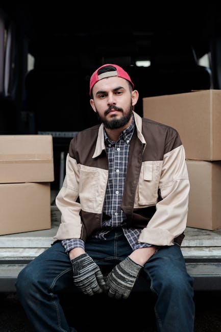 A man dressed in a blue beanie hat, navy and red jacket, black pants, and white sneakers sits on the open rear door of a black moving van during a home relocation process. Inside the van, several card