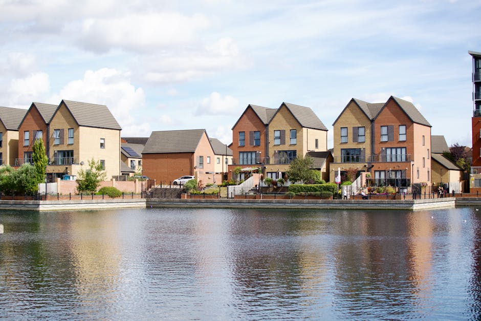 A row of modern, multi-storey residential houses in Roding Valley Estate, Woodford Green, situated along the edge of a calm waterway. The houses feature a variety of brick tones, including beige and reddish-brown, with pitched roofs covered in grey tiles. Each property has a small front garden with landscaping elements such as shrubs, trees, and paved paths. Some houses have external staircases leading to entrances or back patios. The water in the foreground reflects the houses and the partly cloudy sky overhead, creating a serene scene. Based on the page context for estate moves in Roding Valley Estate, this image visually aligns with home relocation and furniture transport services offered by Man with Van Woodford Green, highlighting residential properties typical of area-specific moving logistics.
