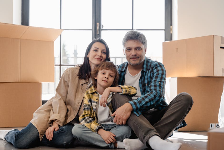 A family of three sitting on the floor inside a new home during a house relocation process, with large cardboard moving boxes stacked nearby. The mother, father, and young son are positioned in front of a large window with natural daylight illuminating the room. The mother, wearing a beige jacket and blue jeans, and the father, in a blue and black checkered shirt and grey trousers, sit close together smiling at the camera. The young boy, dressed in a plaid yellow and black shirt over a white t-shirt, rests his head on his mother’s shoulder, looking calmly at the camera. The scene captures the final stages of packing and preparing for furniture transport, with the boxes likely filled with household items awaiting loading into a moving van. The environment suggests an internal space ready for moving logistics managed by [COMPANY_NAME], specialists in house removals and furniture transport, with natural light highlighting the scene of packing and home relocation activities.