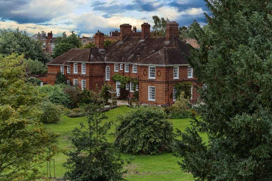 A row of modern, multi-storey residential houses in Roding Valley Estate, Woodford Green, situated along the edge of a calm waterway. The houses feature a variety of brick tones, including beige and reddish-brown, with pitched roofs covered in grey tiles. Each property has a small front garden with landscaping elements such as shrubs, trees, and paved paths. Some houses have external staircases leading to entrances or back patios. The water in the foreground reflects the houses and the partly cloudy sky overhead, creating a serene scene. Based on the page context for estate moves in Roding Valley Estate, this image visually aligns with home relocation and furniture transport services offered by Man with Van Woodford Green, highlighting residential properties typical of area-specific moving logistics.