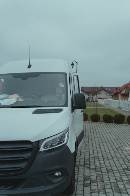 A white delivery van from Man with Van Woodford Green is parked on a cobblestone driveway in a residential area with houses, on a cloudy day. The van's front and side are visible, with the driver inside, partially seen through the windshield. The van has a black side mirror and a black front bumper, with equipment or packing materials placed on the dashboard inside. Behind the vehicle, there is a paved area with neatly trimmed bushes and a white fence bordering the properties. The scene captures the loading or home relocation process outdoors, illustrating a typical house removal setup where furniture and boxes could be unloaded or prepared for transportation. The environment appears quiet and organized, with clear separation between the driveway and the surrounding garden areas, emphasizing professional furniture transport and packing procedures associated with house removals.