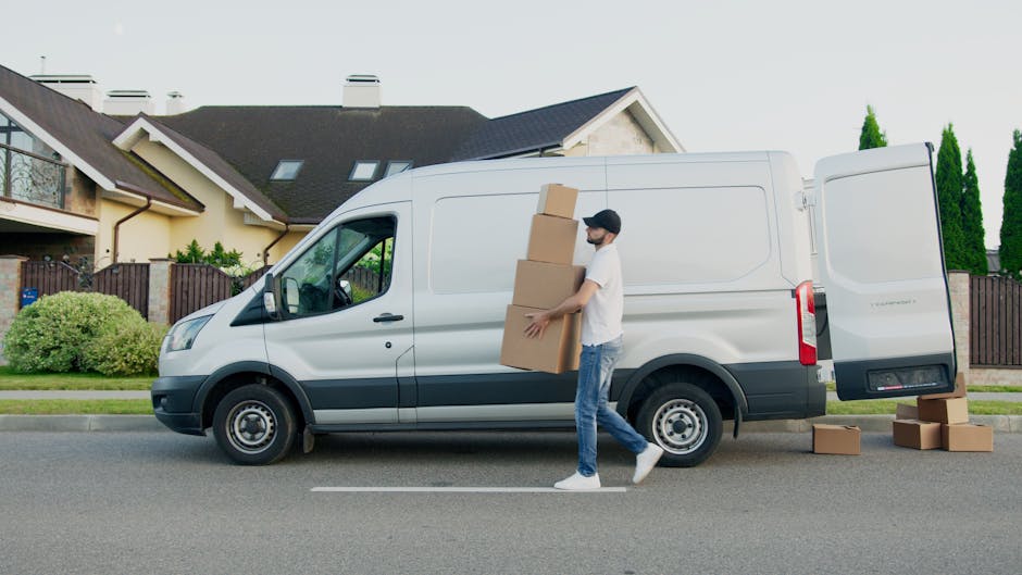 A man dressed in a grey knit beanie, a green padded vest worn over a dark zip-up jacket, and navy trousers, stands outside next to an open loading area of a black moving van. He is holding a small bla