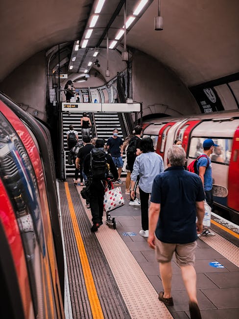 Interior view of a modern underground station with a prominent London Underground roundel sign suspended above the escalator entrance. The station features a glass ceiling with skylights allowing natu