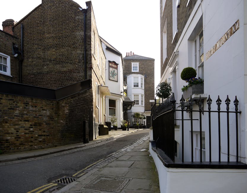 A narrow residential street in Woodford Green with closely packed buildings on both sides. The street is paved with cobblestones and a slightly inclined sidewalk runs along the white-painted house on the right, which features decorative black iron railings and flower boxes with potted plants on the window sills. The neighboring building on the left is constructed from dark brick and has a small, white-framed window. Further down the street, there are additional traditional houses with bay windows and chimneys visible in the background. The scene is lit by natural daylight, highlighting the textures of the brick, paint, and plants. This setting illustrates the typical environment for house removals and furniture transport, where careful planning is required to navigate tight, urban streets during home relocation processes, with [COMPANY_NAME] often handling such logistics as part of their removals services.