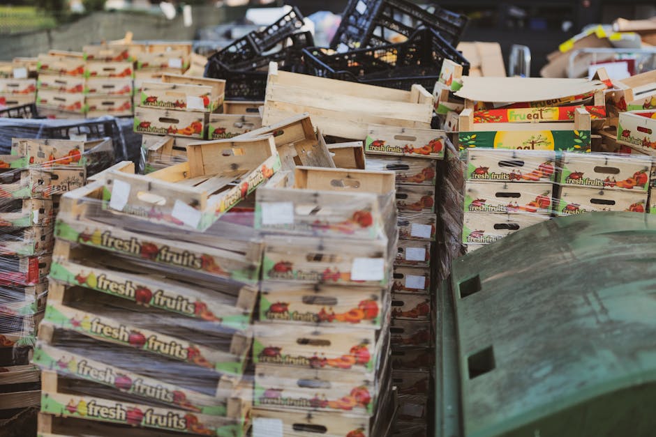 A large collection of wooden and plastic crates filled with cardboard boxes labeled 'fresh fruits' is stacked on the pavement outside a property during a home relocation process. In the foreground, a partially visible green recycling bin is positioned near the crates. Behind the crates, black plastic crates are stacked, and the background shows part of a doorway and a moving van being loaded. The crates contain various items wrapped in paper and plastic, indicating packing materials used for furniture and household items during a move. The scene is lit by natural daylight, capturing the organized packing process associated with professional removals, such as those offered by Man with Van Woodford Green, in accordance with local waste disposal rules for bulky items in Woodford Green.