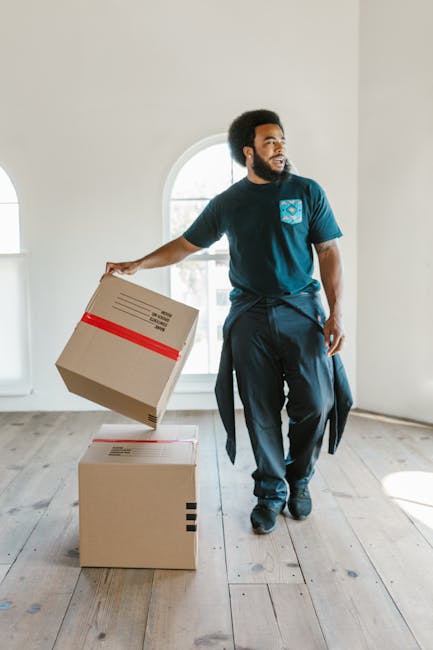 A man with dark skin and curly black hair, dressed in a navy blue t-shirt with a logo on the chest and dark trousers, is inside a bright, empty room with wooden flooring and white walls. He is holding a cardboard moving box in his right hand, which is partially lifted, and is standing next to two more cardboard boxes, one on top of the other, positioned on the floor near a large arched window that allows natural light to fill the space. The room appears to be in the process of home relocation, with the man likely engaged in packing or furniture transport as part of a same-day removals service. The setting suggests the early stages of a house move, with minimal furnishings and open space, highlighting the logistics involved in packing and preparing household items for transport, supported by the professional services of [COMPANY_NAME], such as House removals and moving solutions.