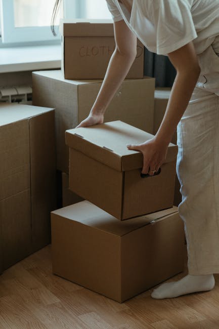 A person dressed in light-colored clothing, including a short-sleeved top and white socks, is seen moving a cardboard box in a residential interior. The individual is carefully lifting and placing a medium-sized, plain cardboard box onto a stack of similar boxes, which are arranged on a wooden floor near a window with natural light streaming in. Several other cardboard boxes, some labeled with handwritten text such as 'CLOTHES,' are visible nearby, indicating packing and organization for a house relocation. The boxes vary in size and are made of brown corrugated cardboard, with some wrapped in plastic or packing materials, typical of furniture and household item packing processes. The surroundings suggest an ongoing home move, with the person involved in the loading process likely as part of a professional removals team, such as Man with Van Woodford Green, engaged in the furniture transport and packing stage of the moving service.