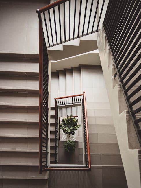 A view from above of a multi-flight staircase in a residential building with beige tiled steps and dark wooden handrails. The stairwell features two small potted plants placed on a platform at the landing, with one on a decorative ceramic plate. The surrounding walls are painted in neutral tones, and the staircase is lit by natural light coming from the upper floors. The image captures the orderly and clean environment, demonstrating the internal layout of a staircase-only flat complex on Snakes Lane. This setting reflects typical residential moving environments where careful handling of furniture and packing materials is essential during home relocations. Man with Van Woodford Green, known for professional removals services, often manages the safe transport of furniture and boxes through such staircase areas during house removals and furniture transport in the Woodford Green area.