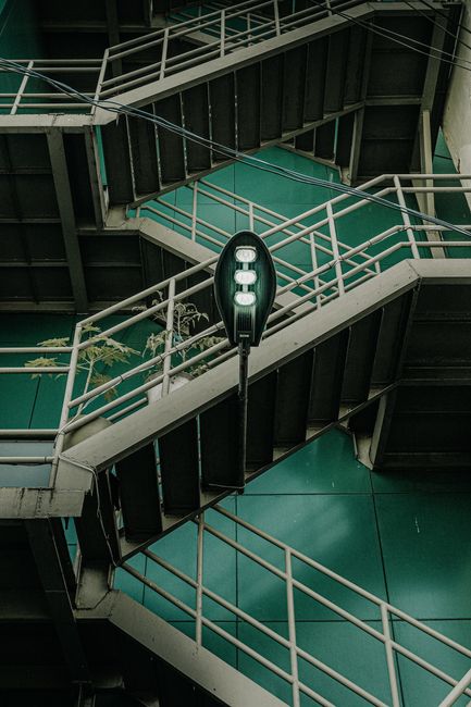 A multi-level outdoor metal staircase with white handrails and dark steps, positioned against a green wall. The stairs zigzag across the frame, with a black streetlamp hanging between different sections of the staircase. The environment is illuminated with natural light, and a small potted plant is visible on the left side of the image. This setting illustrates a typical exterior staircase often found in residential or urban buildings, relevant to home relocation and furniture transport processes, as seen in house removals services provided by Man with Van Woodford Green.