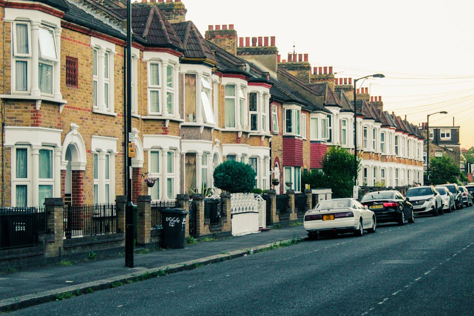 A row of terraced houses on a residential street with brick facades and bay windows, some open with visible curtains. In the foreground, a sidewalk runs parallel to the street, with a black iron fence and small front gardens or yards. Several parked cars, including sedans and hatchbacks, line the curb, with a white car closest to the camera. The street is wide and appears to be quiet, with street lamps and overhead utility wires visible. The lighting suggests late afternoon or early evening. The scene depicts an urban residential area suitable for home relocation and furniture transport, with the environment prepared for moving logistics and packing activities. Man with Van Woodford Green occasionally services moves across streets like this, aiding in efficient loading and unloading processes during house removals.
