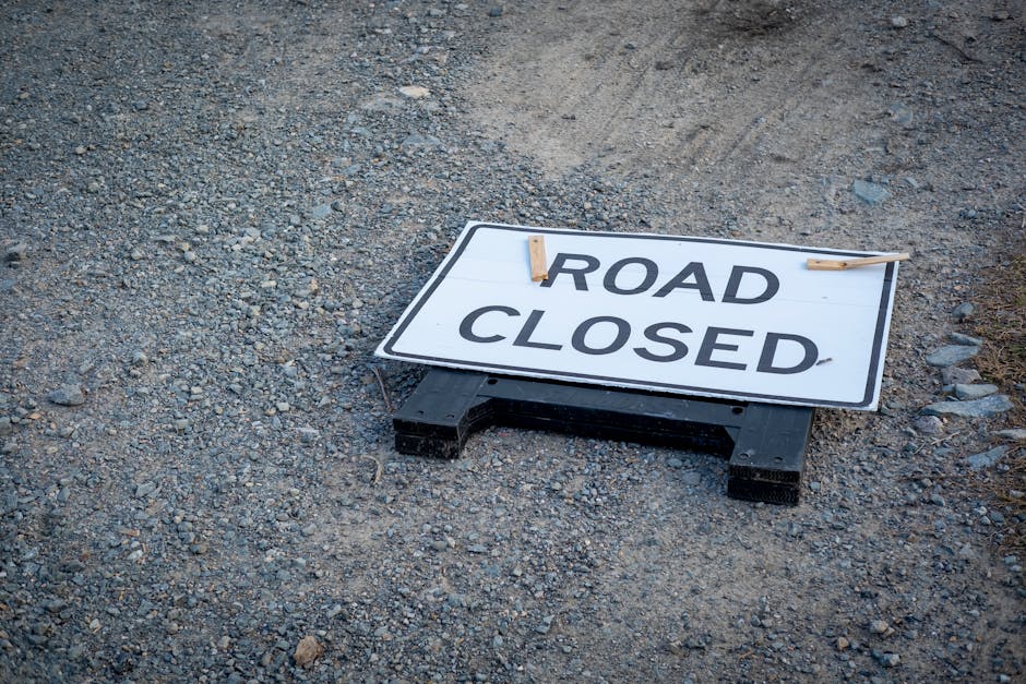 A rectangular white road closure sign with bold black lettering reading 'ROAD CLOSED' is lying down on a gravel surface, supported by a black plastic base at the bottom. The sign is positioned near the edge of a paved or tarmac area, with its top edge slightly tilted. Two wooden clothespins are clipped onto the upper corners of the sign, holding it in place. The surrounding area consists of loose gravel and small stones with no other objects or vehicles visible. The scene appears to be outdoors, possibly on a construction site or street undergoing roadworks, relevant to street closure permits and managing access for house removals and furniture transport. This setting supports the context of traffic restrictions impacting local relocations, as documented by Man with Van Woodford Green, which specializes in removals services.