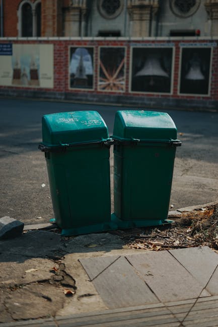 Two green wheelie bins with closed lids positioned side by side on a pavement near the curb outside a property, with one bin slightly taller than the other. The bins are placed on a concrete surface, with some dirt and broken pavement debris around their bases. Behind the bins, there is a street with an asphalt road, and a building with large windows and decorative brickwork visible in the background. The scene appears to be outside a residential or commercial property, and the bins are likely used for waste or recycling collection, part of the local waste management services. This image relates to house removals and moving services, such as clearing waste or bulky items during a home relocation, which [COMPANY_NAME] offers as part of their removals process.