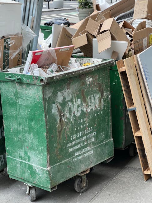 A man with curly hair, dressed in a black t-shirt and striped shorts, is leaning over and examining a large white skip filled with various building materials, including wooden planks and tiles, situated on a paved street in an urban area. The skip is positioned next to a metal fence and a sidewalk, with some greenery visible on the right side of the image. In the background, there are residential and commercial buildings, a few pedestrians, and several blue and white signs mounted on poles. The scene depicts a waste clearance or home disposal activity related to a house removal or renovation project, with the skip used for disposing of bulky waste materials. This image illustrates the process of loading and managing waste as part of a home relocation, with a focus on practical removal solutions provided by companies like Man with Van Woodford Green, supporting efficient house removals and furniture transport services.
